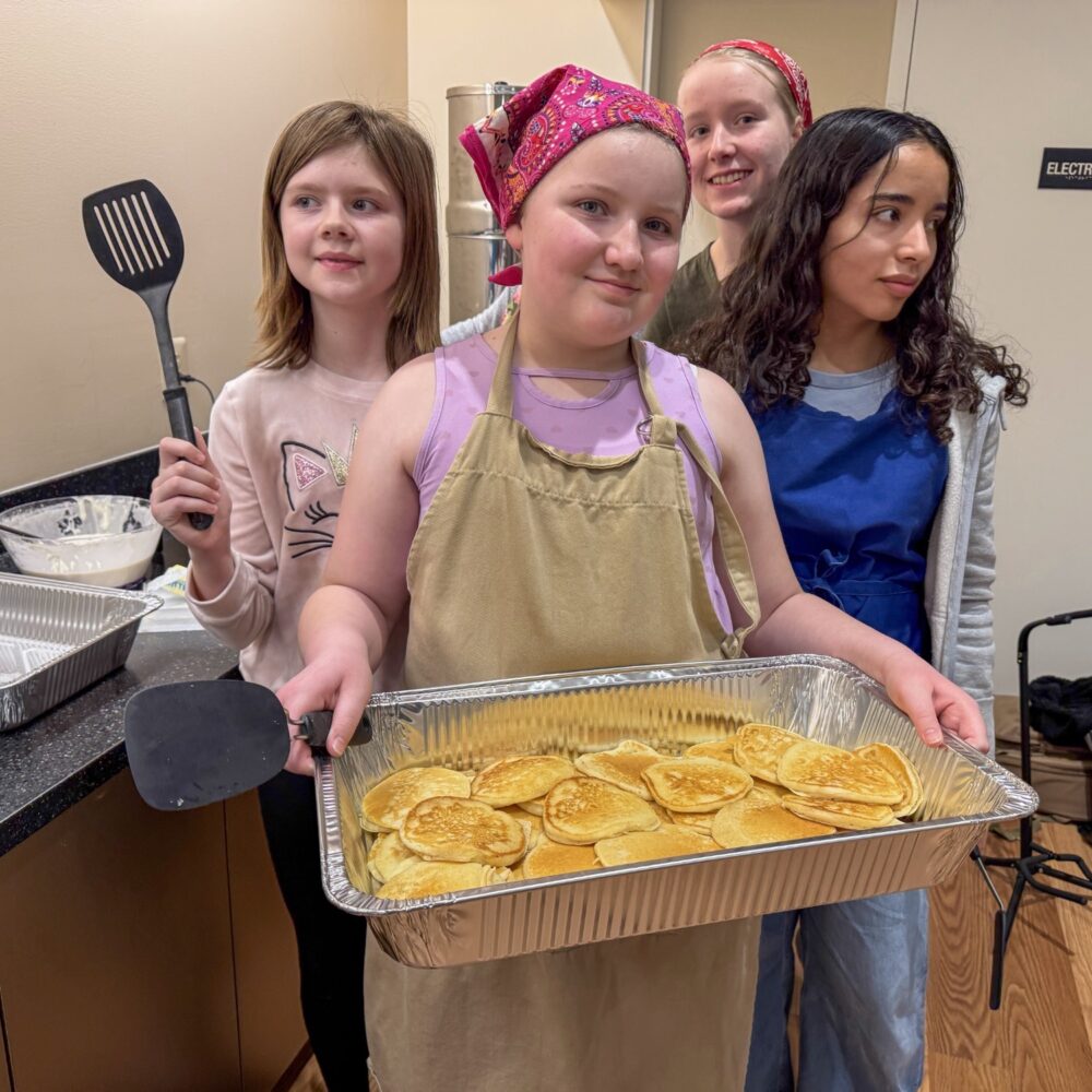 Group of young people in aprons working together in the congregation kitchen, the one in front holding up a foil tray of food they've prepared