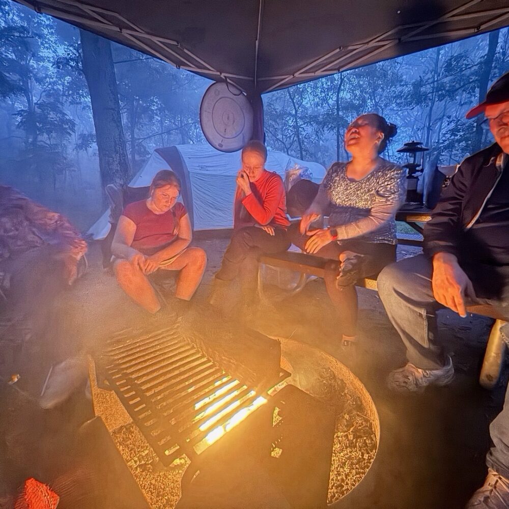 Group of people gathered around an outdoor fire pit at twilight, with a deep blue sky overhead and warm light from the flames