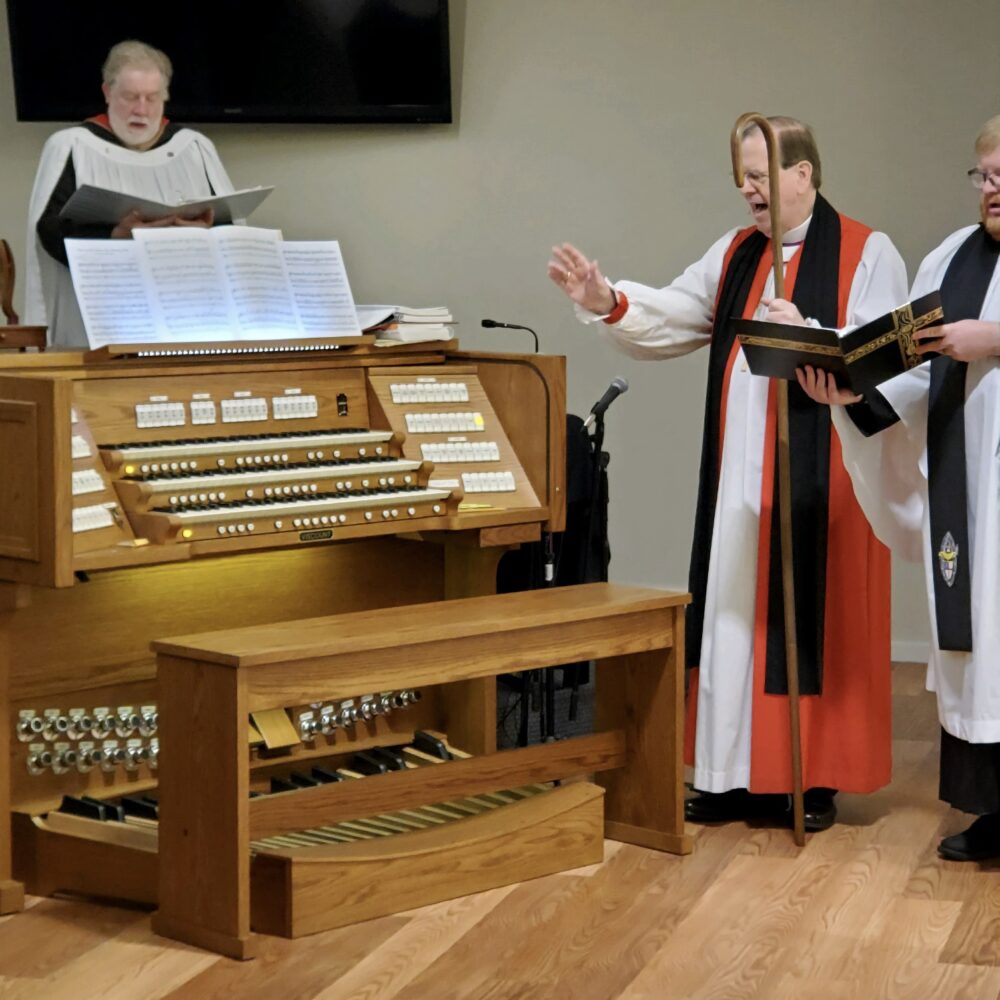 Three clergy members standing at the organ console during a service, one playing the organ and two others reading from a service book beside it